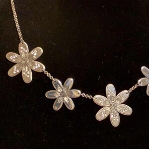 Może przedstawiać: Srebrny naszyjnik z zawieszkami w kształcie stokrotek. Każdy kwiat ma spiralny środek i jest ozdobiony małymi, błyszczącymi akcentami. Naszyjnik jest prezentowany na ciemnym tle, podkreślając szczeg&oacute;ły.