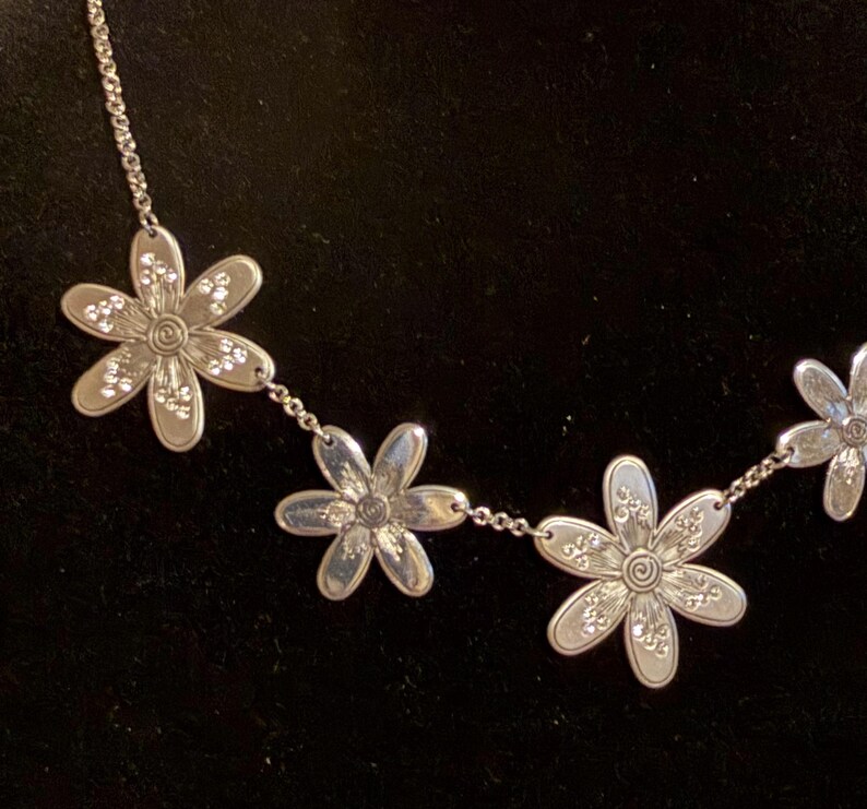 Może przedstawiać: Srebrny naszyjnik z zawieszkami w kształcie stokrotek. Każdy kwiat ma spiralny środek i jest ozdobiony małymi, błyszczącymi akcentami. Naszyjnik jest prezentowany na ciemnym tle, podkreślając szczeg&oacute;ły.