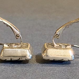 Puede incluir: Un par de pendientes plateados. Cada pendiente presenta una base cuadrada y texturizada con un patr&oacute;n decorativo y un alambre para la oreja curvado y con bisagras. Los pendientes se muestran sobre un fondo gris.