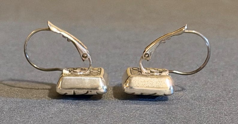 Puede incluir: Un par de pendientes plateados. Cada pendiente presenta una base cuadrada y texturizada con un patr&oacute;n decorativo y un alambre para la oreja curvado y con bisagras. Los pendientes se muestran sobre un fondo gris.