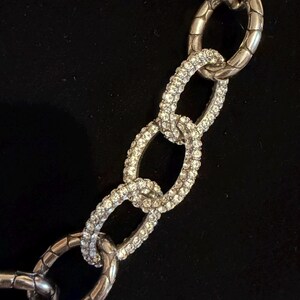 Puede incluir: Primer plano de un collar de cadena plateada con eslabones ovalados grandes. Algunos eslabones est&aacute;n incrustados con cristales transparentes brillantes, mientras que otros tienen un dise&ntilde;o texturizado y acanalado. El collar est&aacute; sobre un fondo negro.