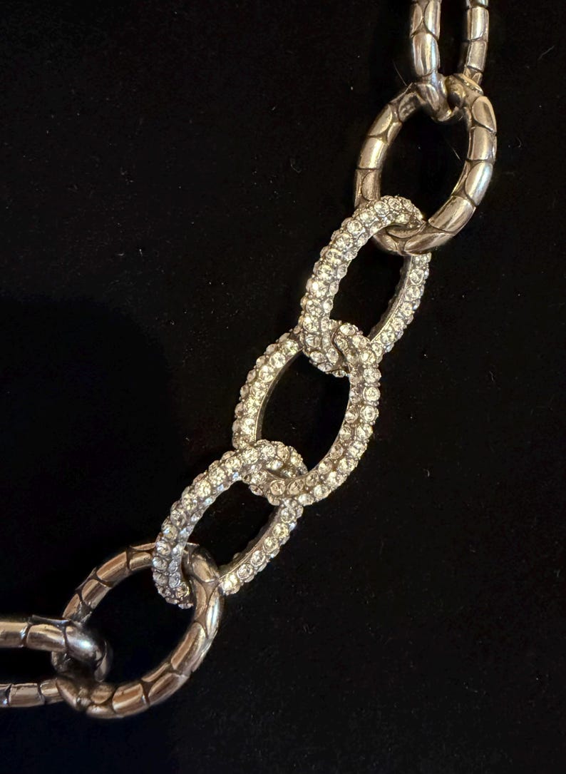 Puede incluir: Primer plano de un collar de cadena plateada con eslabones ovalados grandes. Algunos eslabones est&aacute;n incrustados con cristales transparentes brillantes, mientras que otros tienen un dise&ntilde;o texturizado y acanalado. El collar est&aacute; sobre un fondo negro.