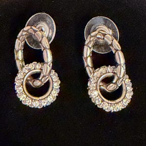 Puede incluir: Un par de pendientes plateados. Cada pendiente presenta un eslab&oacute;n ovalado texturizado conectado a un eslab&oacute;n circular m&aacute;s peque&ntilde;o incrustado con peque&ntilde;os cristales transparentes. Los pendientes tienen cierres de poste.