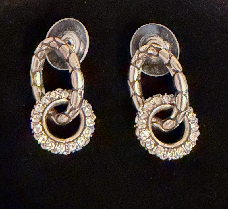 Puede incluir: Un par de pendientes plateados. Cada pendiente presenta un eslab&oacute;n ovalado texturizado conectado a un eslab&oacute;n circular m&aacute;s peque&ntilde;o incrustado con peque&ntilde;os cristales transparentes. Los pendientes tienen cierres de poste.