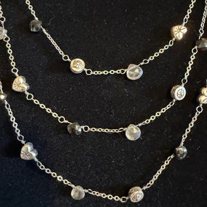 May include: A three-strand necklace with silver chains and decorative charms. The necklace features heart-shaped and round charms, alongside clear and dark grey faceted beads. The chains are set against a black backdrop.