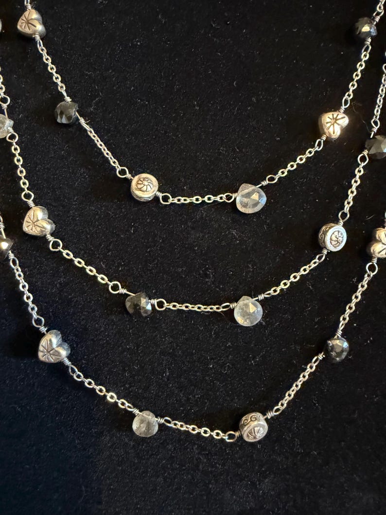 May include: A three-strand necklace with silver chains and decorative charms. The necklace features heart-shaped and round charms, alongside clear and dark grey faceted beads. The chains are set against a black backdrop.