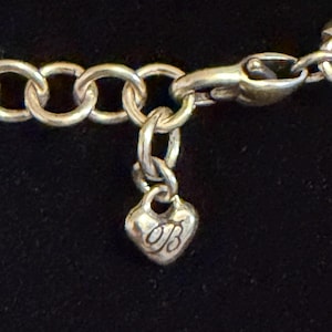 May include: Silver necklace with a heart-shaped charm. The charm features a cursive "OB" design. The necklace has a lobster clasp and a series of circular links. The background is a solid black.