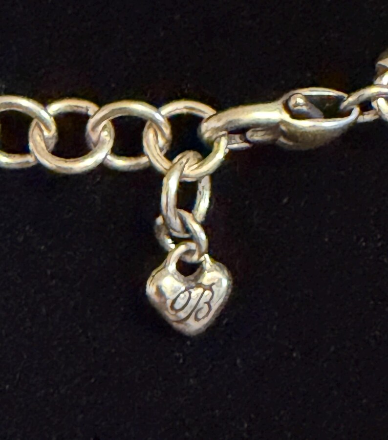 May include: Silver necklace with a heart-shaped charm. The charm features a cursive "OB" design. The necklace has a lobster clasp and a series of circular links. The background is a solid black.