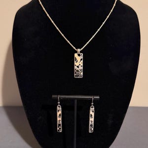 K&ouml;nnte beinhalten: Ein silbernes Halsketten- und Ohrring-Set. Der Anh&auml;nger und die Ohrringe haben eine rechteckige Form mit einem schwarz-wei&szlig;en Blumendesign. Der Schmuck wird auf einer schwarzen Samtb&uuml;ste und einem St&auml;nder pr&auml;sentiert.