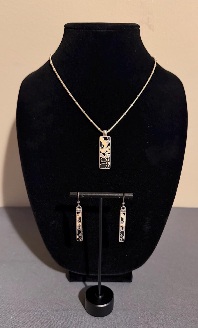 K&ouml;nnte beinhalten: Ein silbernes Halsketten- und Ohrring-Set. Der Anh&auml;nger und die Ohrringe haben eine rechteckige Form mit einem schwarz-wei&szlig;en Blumendesign. Der Schmuck wird auf einer schwarzen Samtb&uuml;ste und einem St&auml;nder pr&auml;sentiert.