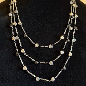 May include: A three-strand silver necklace with clear and dark grey beads. The necklace features small heart and round-shaped charms. The background is black.