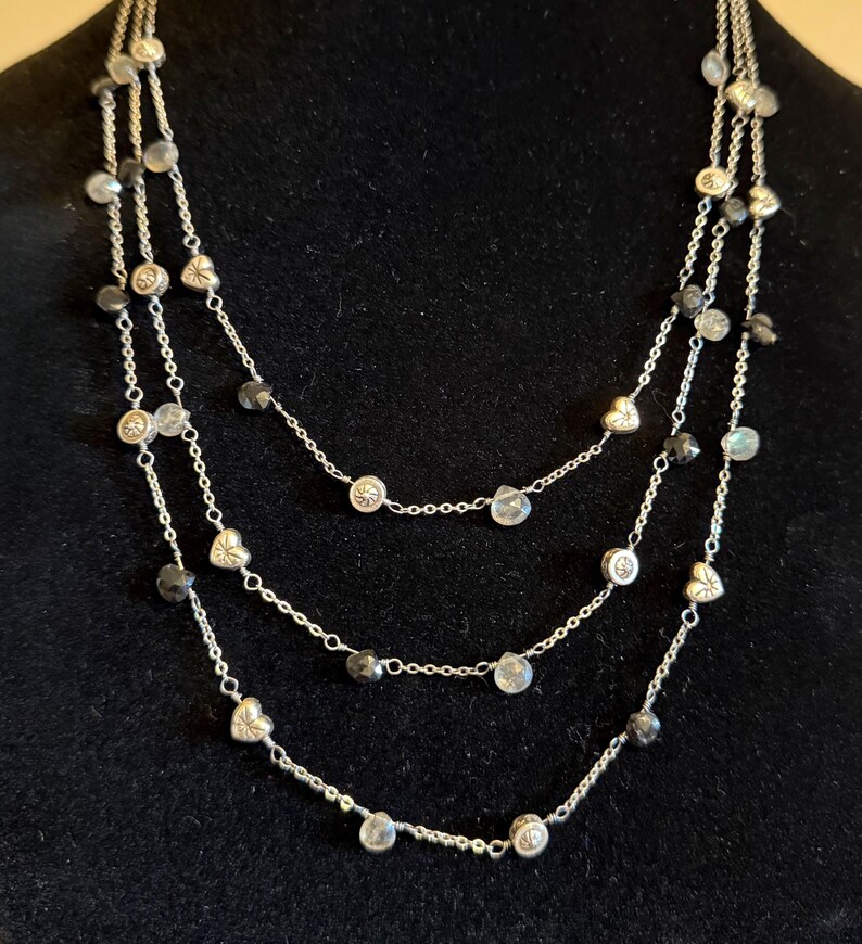 May include: A three-strand silver necklace with clear and dark grey beads. The necklace features small heart and round-shaped charms. The background is black.