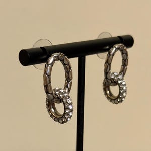 Puede incluir: Un par de pendientes plateados con un dise&ntilde;o de doble lazo. El lazo superior tiene un patr&oacute;n texturizado, mientras que el lazo inferior est&aacute; incrustado con peque&ntilde;os cristales transparentes. Los pendientes se exhiben en un soporte negro.