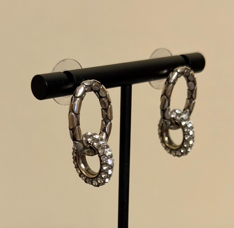 Puede incluir: Un par de pendientes plateados con un dise&ntilde;o de doble lazo. El lazo superior tiene un patr&oacute;n texturizado, mientras que el lazo inferior est&aacute; incrustado con peque&ntilde;os cristales transparentes. Los pendientes se exhiben en un soporte negro.