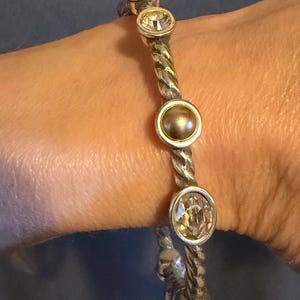 May include: Silver-toned bracelet with a twisted rope design. The bracelet features four circular, bezel-set gemstones: three clear, faceted stones and one pearl-like stone. The bracelet is worn on a wrist.