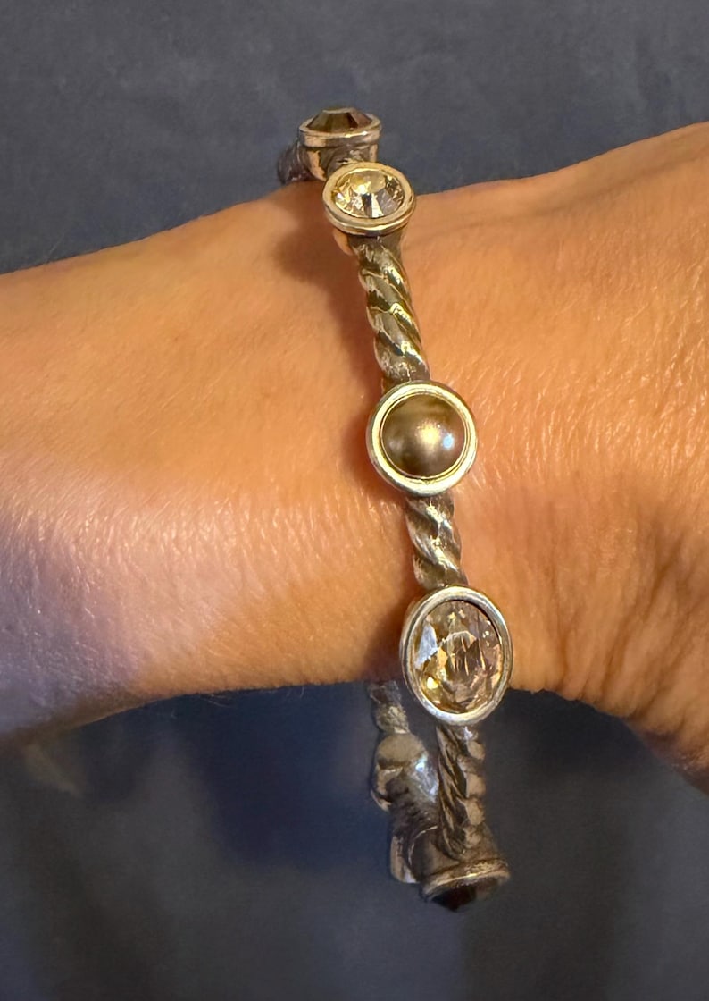 May include: Silver-toned bracelet with a twisted rope design. The bracelet features four circular, bezel-set gemstones: three clear, faceted stones and one pearl-like stone. The bracelet is worn on a wrist.