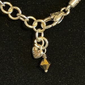 May include: Close-up of a silver-toned necklace with a heart-shaped charm and a faceted, dark-colored bead. The necklace features a chain with large, round links and a lobster clasp. The heart charm has an engraved design.