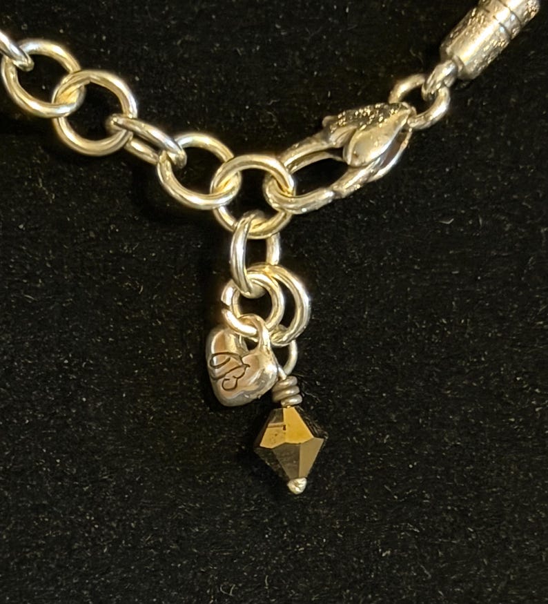 May include: Close-up of a silver-toned necklace with a heart-shaped charm and a faceted, dark-colored bead. The necklace features a chain with large, round links and a lobster clasp. The heart charm has an engraved design.