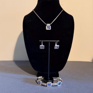 Puede incluir: Un conjunto de joyas plateadas que incluye un collar, pendientes y pulsera. Cada pieza tiene una gema azul cuadrada en el centro. El collar y los pendientes se exhiben en un soporte negro, mientras que la pulsera descansa debajo.