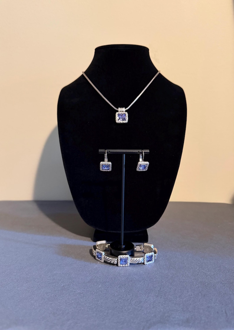Puede incluir: Un conjunto de joyas plateadas que incluye un collar, pendientes y pulsera. Cada pieza tiene una gema azul cuadrada en el centro. El collar y los pendientes se exhiben en un soporte negro, mientras que la pulsera descansa debajo.