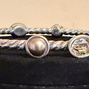 May include: Two silver-toned bangles with a twisted rope design. One bangle features a central, round, light brown gem, and the other has a series of oval-shaped, light brown gems. Additional small, round gems are also present.