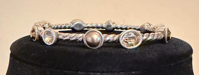 May include: Two silver-toned bangles with a twisted rope design. One bangle features a central, round, light brown gem, and the other has a series of oval-shaped, light brown gems. Additional small, round gems are also present.