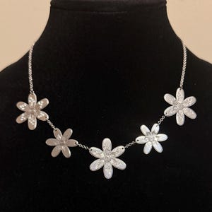 Może przedstawiać: Srebrny naszyjnik z pięcioma wisiorkami w kształcie stokrotek. Każdy kwiat ma szczeg&oacute;łowe wzory płatk&oacute;w i środka, połączone delikatnym łańcuszkiem. Naszyjnik jest prezentowany na czarnym manekinie.