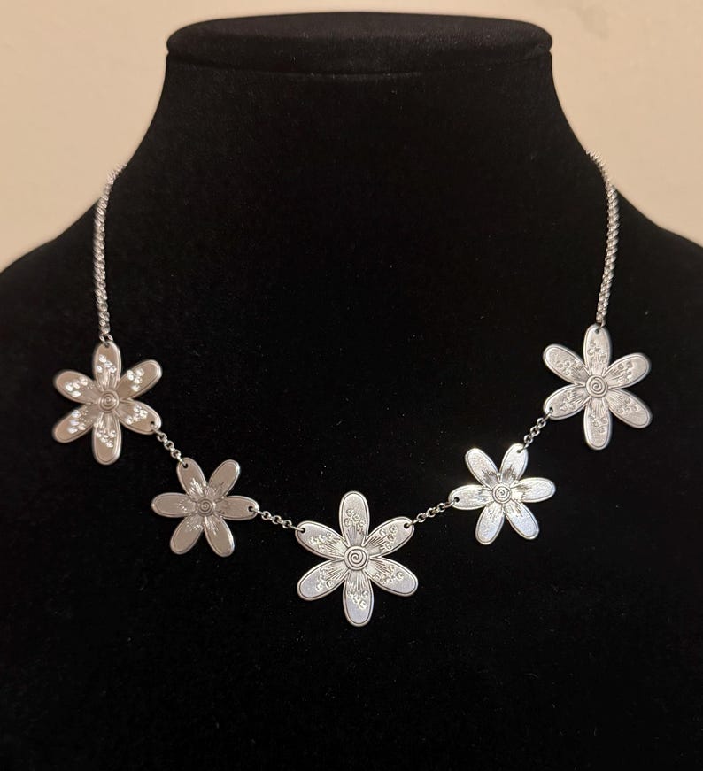 Może przedstawiać: Srebrny naszyjnik z pięcioma wisiorkami w kształcie stokrotek. Każdy kwiat ma szczeg&oacute;łowe wzory płatk&oacute;w i środka, połączone delikatnym łańcuszkiem. Naszyjnik jest prezentowany na czarnym manekinie.