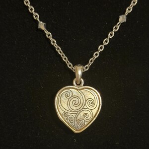 May include: Silver-toned necklace with a heart-shaped pendant. The pendant has a gold-colored center with a swirling design and a silver-colored border. The chain is made of small, linked pieces.