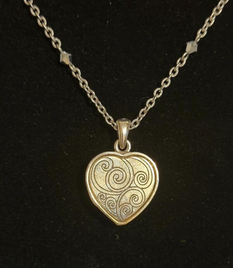 May include: Silver-toned necklace with a heart-shaped pendant. The pendant has a gold-colored center with a swirling design and a silver-colored border. The chain is made of small, linked pieces.