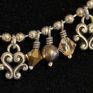 May include: Silver-toned necklace with decorative heart-shaped charms, faceted crystals, and a dark gray pearl-like bead. The necklace has a beaded chain and is set against a dark background.