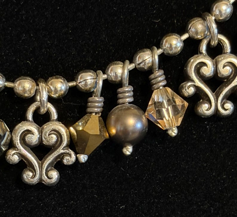 May include: Silver-toned necklace with decorative heart-shaped charms, faceted crystals, and a dark gray pearl-like bead. The necklace has a beaded chain and is set against a dark background.