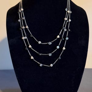 May include: A three-strand necklace with silver chains and various embellishments. The necklace features clear and black beads, along with heart-shaped and round silver accents. The jewelry is displayed on a black mannequin.