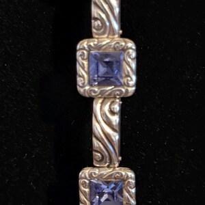 Puede incluir: Pulsera de plata con piedras preciosas azules cuadradas engastadas en marcos cuadrados ornamentados. Los eslabones de la pulsera tienen un dise&ntilde;o arremolinado y texturizado. El fondo es de color oscuro y s&oacute;lido.