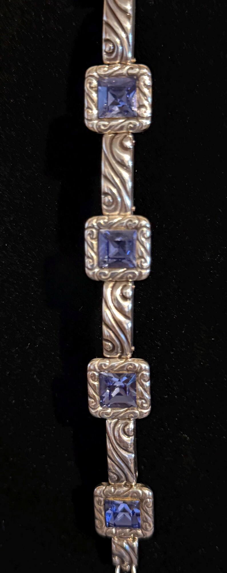 Puede incluir: Pulsera de plata con piedras preciosas azules cuadradas engastadas en marcos cuadrados ornamentados. Los eslabones de la pulsera tienen un dise&ntilde;o arremolinado y texturizado. El fondo es de color oscuro y s&oacute;lido.