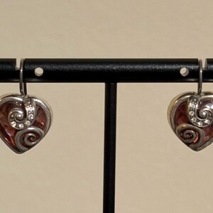 May include: A pair of heart-shaped earrings with a silver-tone metal frame. The earrings feature a reddish-brown center with a swirling design and small clear crystals. The earrings are displayed on a black stand.
