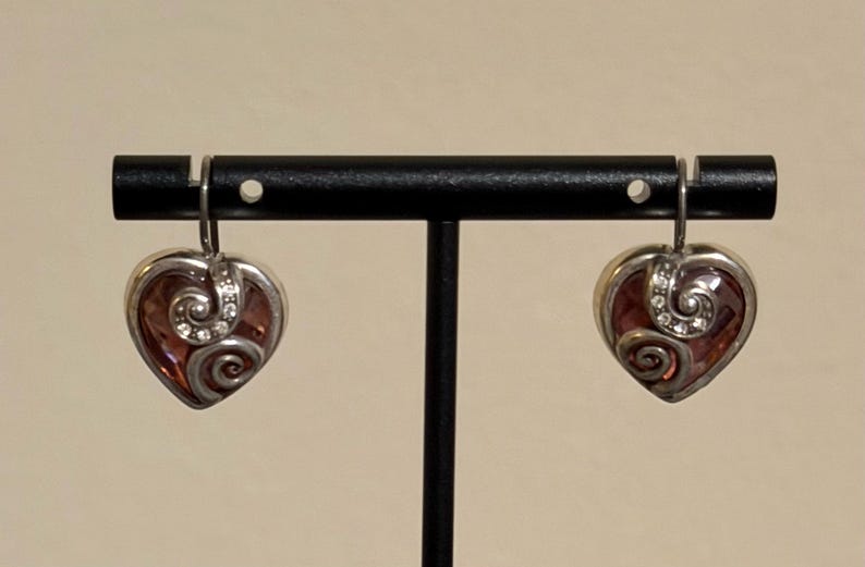 May include: A pair of heart-shaped earrings with a silver-tone metal frame. The earrings feature a reddish-brown center with a swirling design and small clear crystals. The earrings are displayed on a black stand.