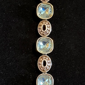 May include: Silver bracelet with oval, pale blue gemstones set in decorative silver frames. The bracelet has a chain extension and a heart-shaped clasp. The background is black.