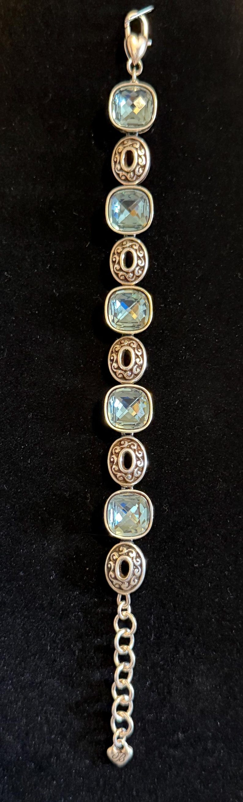 May include: Silver bracelet with oval, pale blue gemstones set in decorative silver frames. The bracelet has a chain extension and a heart-shaped clasp. The background is black.