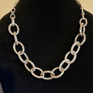 Puede incluir: Collar de cadena plateada con eslabones ovalados texturizados. Una secci&oacute;n del collar presenta eslabones ovalados incrustados con peque&ntilde;os cristales transparentes. El collar se exhibe sobre una superficie negra.