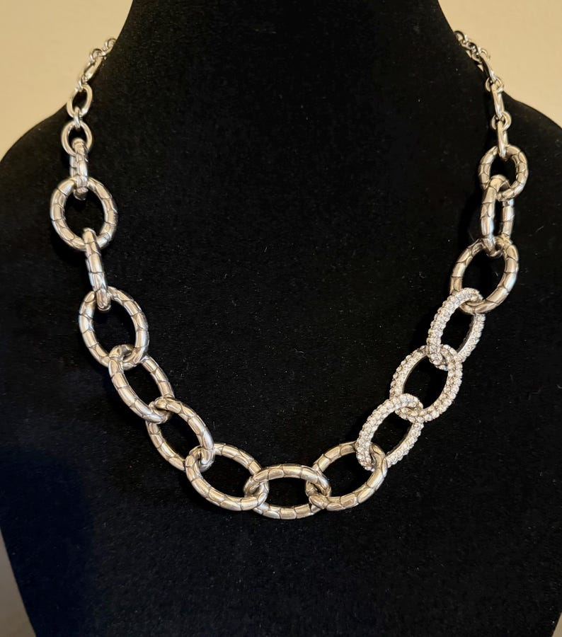 Puede incluir: Collar de cadena plateada con eslabones ovalados texturizados. Una secci&oacute;n del collar presenta eslabones ovalados incrustados con peque&ntilde;os cristales transparentes. El collar se exhibe sobre una superficie negra.