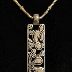 K&ouml;nnte beinhalten: Silberfarbene Anh&auml;ngerkette mit einem rechteckigen Anh&auml;nger mit floralem Design. Der Anh&auml;nger h&auml;ngt an einer filigranen Kette. Die Halskette wird vor schwarzem Hintergrund pr&auml;sentiert.