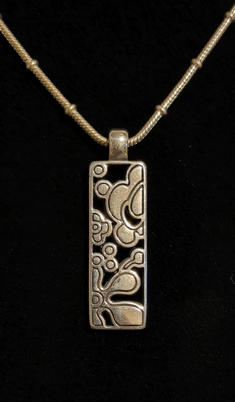 K&ouml;nnte beinhalten: Silberfarbene Anh&auml;ngerkette mit einem rechteckigen Anh&auml;nger mit floralem Design. Der Anh&auml;nger h&auml;ngt an einer filigranen Kette. Die Halskette wird vor schwarzem Hintergrund pr&auml;sentiert.