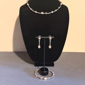 May include: A silver-toned jewelry set displayed on a black mannequin. The set includes a necklace, earrings, and a bracelet. The necklace and bracelet feature oval-shaped embellishments. The earrings are dangling. The jewelry is displayed on a gray surface.