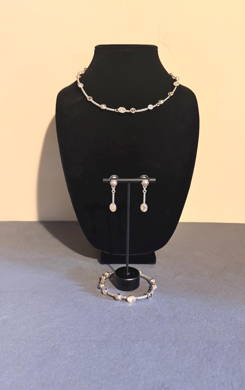 May include: A silver-toned jewelry set displayed on a black mannequin. The set includes a necklace, earrings, and a bracelet. The necklace and bracelet feature oval-shaped embellishments. The earrings are dangling. The jewelry is displayed on a gray surface.