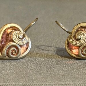 May include: A pair of heart-shaped earrings with a silver-tone metal frame. The earrings feature a pinkish-brown center with a swirl design and small clear stones. The earrings have a hook closure.