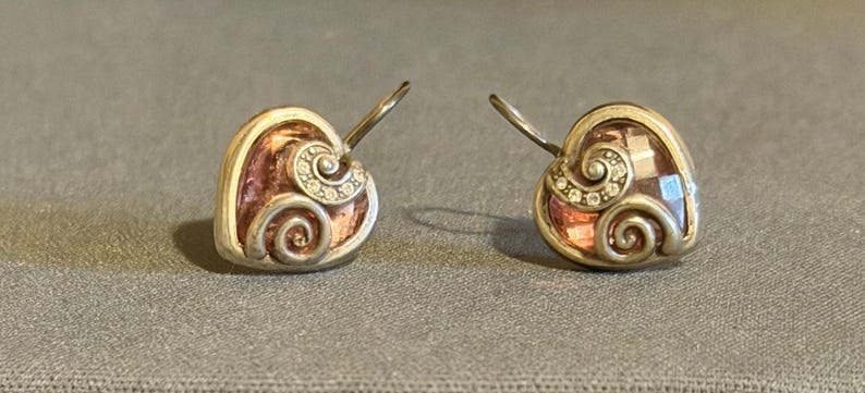 May include: A pair of heart-shaped earrings with a silver-tone metal frame. The earrings feature a pinkish-brown center with a swirl design and small clear stones. The earrings have a hook closure.