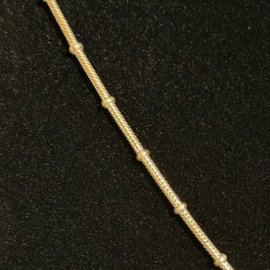 K&ouml;nnte beinhalten: Eine goldfarbene Halskette mit einer strukturierten, seilartigen Kette. Die Kette ist mit gleichm&auml;&szlig;ig verteilten, kleinen, runden Perlen verziert. Die Halskette wird vor einem dunklen Hintergrund pr&auml;sentiert.