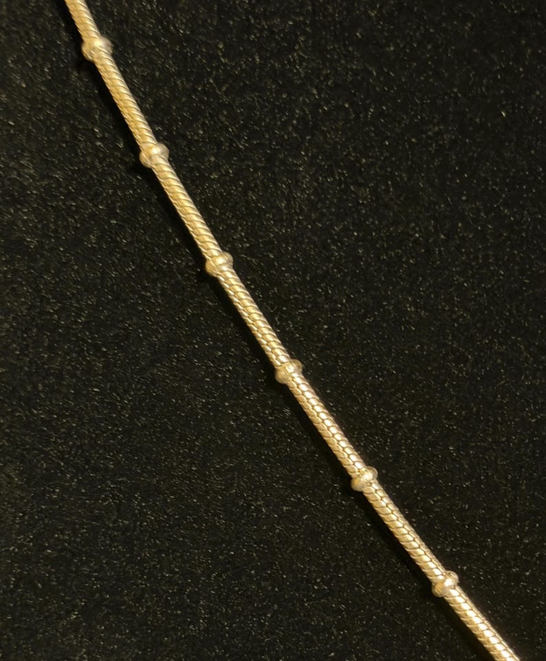 K&ouml;nnte beinhalten: Eine goldfarbene Halskette mit einer strukturierten, seilartigen Kette. Die Kette ist mit gleichm&auml;&szlig;ig verteilten, kleinen, runden Perlen verziert. Die Halskette wird vor einem dunklen Hintergrund pr&auml;sentiert.