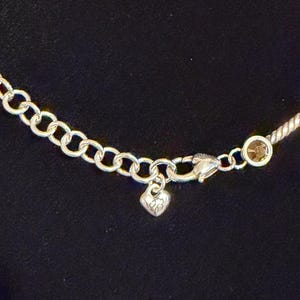 May include: Silver-toned necklace with a delicate chain and a heart-shaped charm. The necklace features small, round, gold-colored accents and a lobster clasp closure. The background is a solid black.